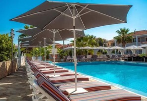 Un área junto a la piscina con sillones de descanso y sombrillas, una piscina azul cristalina y un camino de piedra.