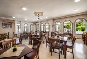 A spacious dining area with multiple tables and chairs, large windows with arched tops, and a ceiling fan. - Anamar Zante Hotel (Zakynthos)