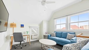 A modern room with a blue sofa, a small round table, a grey office chair, a ceiling fan, and a large window with a view of buildings. - The Melrose, 2B - Double Bed & Bath: Romantic Geta (Neptune Township)