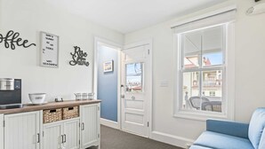 A modern living room with a blue sofa, a white cabinet with baskets, a coffee station, and a view of the outside through a large window. - The Melrose, 2B - Double Bed & Bath: Romantic Geta (Neptune Township)