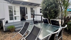 Een buitenterras met een tafel met glazen blad en metalen stoelen, een kleine potplant en een wit gebouw met een schuifdeur op de achtergrond.