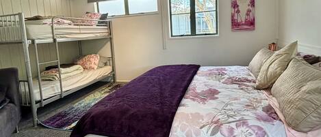 Une chambre avec des lits superposés, un grand lit recouvert d’une couette à motifs floraux, un plaid violet et une fenêtre donnant sur des palmiers.