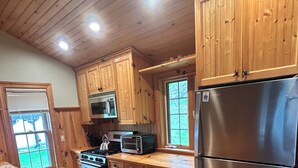 A kitchen with wooden cabinets, a stainless steel refrigerator, a microwave, a stove, and a window with blinds. - Newfound Lake Bristol, NH Lakefront Cottage (Bristol)