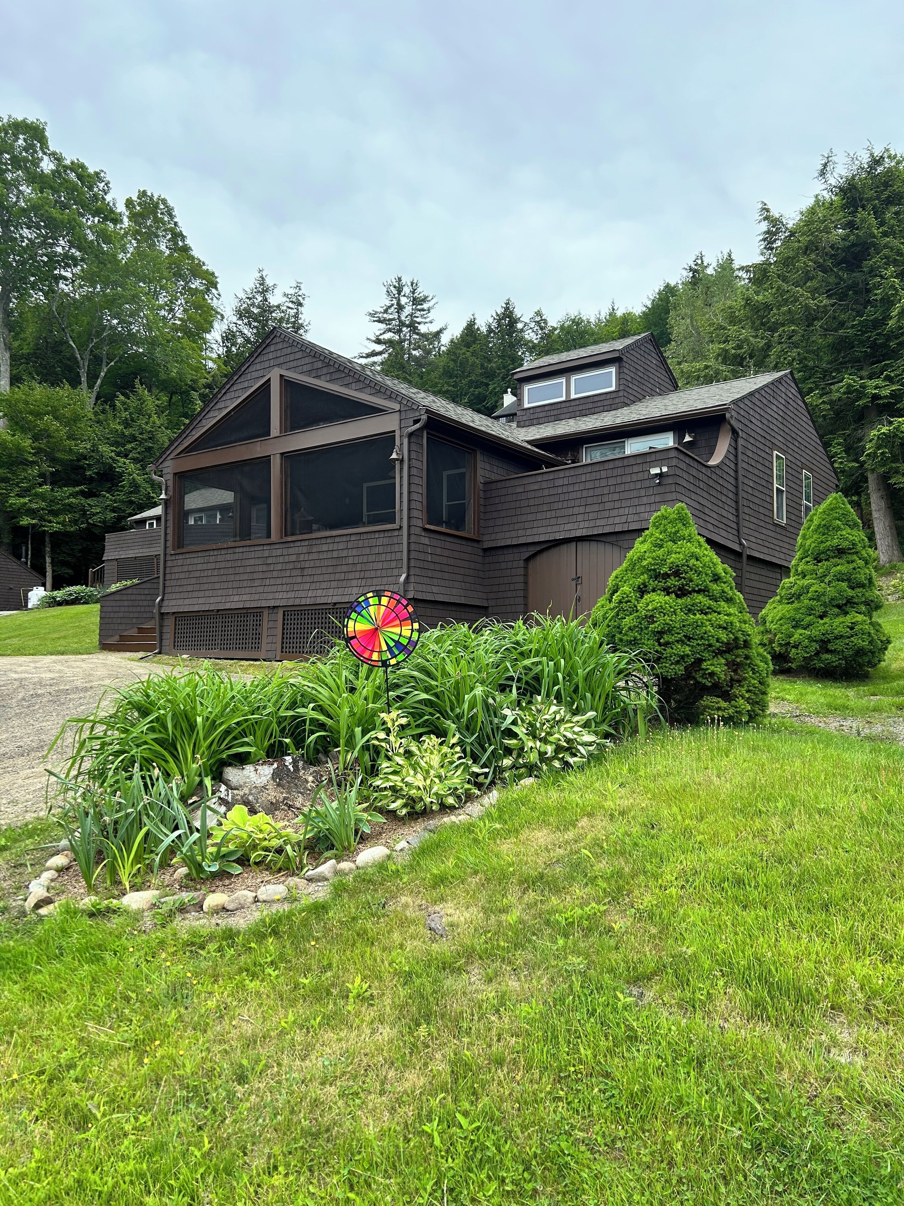A modern two-story house with a dark brown exterior, large windows, and a well-maintained garden with various plants and a colorful windmill.