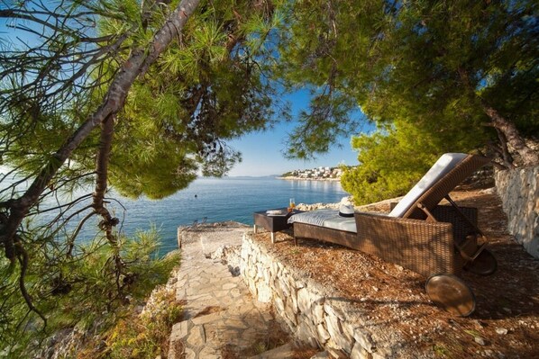 Relax by the sea with a wicker chair on a stone ledge, surrounded by pine trees and a stone wall.