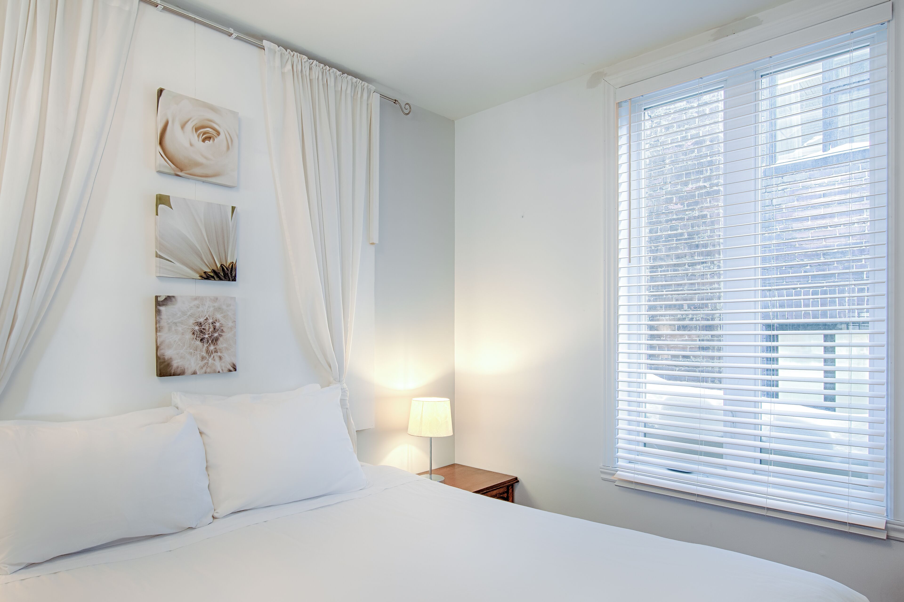A bedroom with a bed, white curtains, a nightstand with a lamp, and a window with blinds.