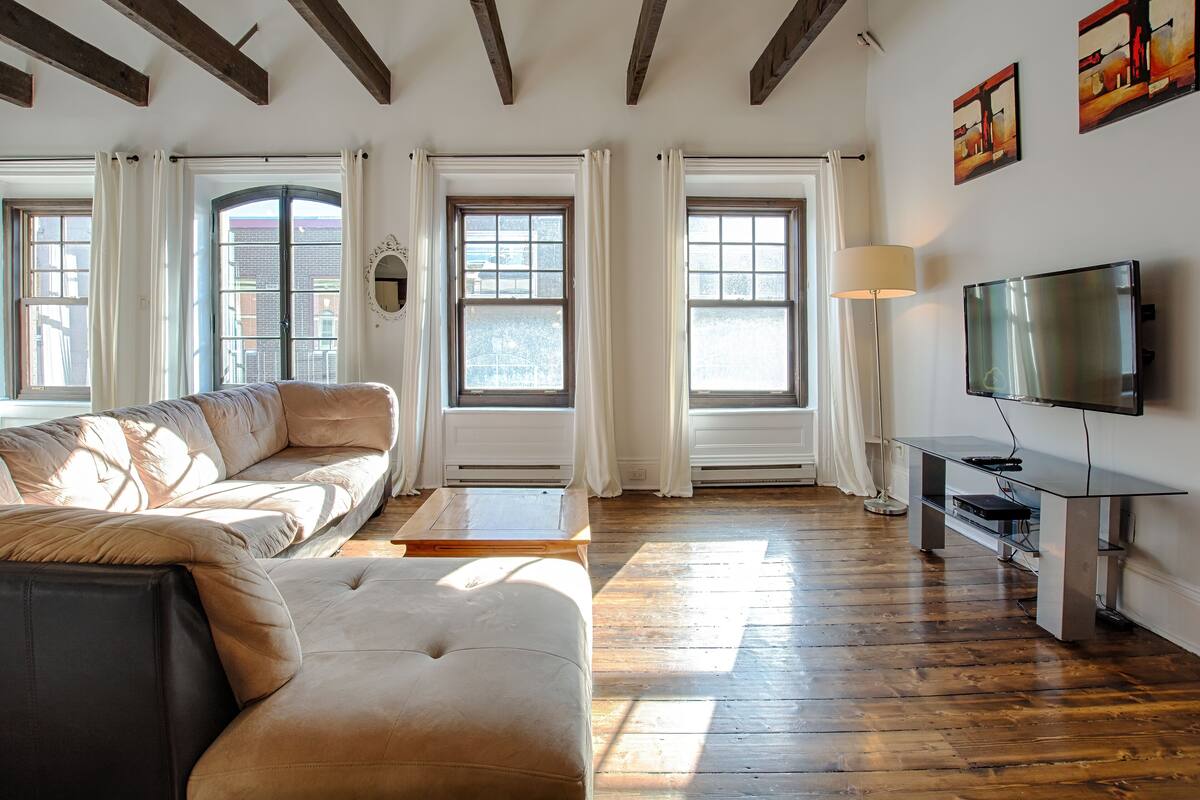 A living room with a sofa, ottoman, wooden coffee table, and a TV on a stand. There are large windows with curtains, a floor lamp, and artwork on the wall.
