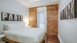 Een slaapkamer met een bed, nachtkastje, lamp, bakstenen muur en een deur.