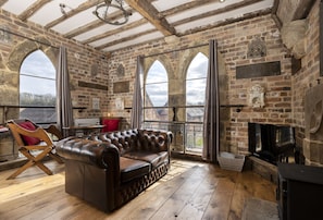 A rustic room with exposed brick walls, a leather sofa, wooden flooring, and large arched windows. - Exposed timbers, a feature stone fireplace, flagstone and oak floors – The Chapel has a wealth of pe (Bromsgrove)