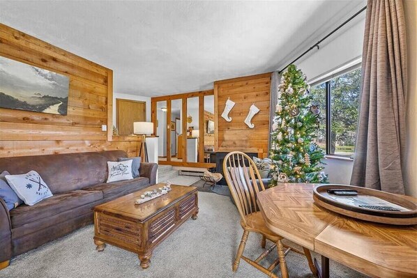 A cozy living room with a wooden wall, a sofa, a coffee table, a dining table with chairs, and a Christmas tree.