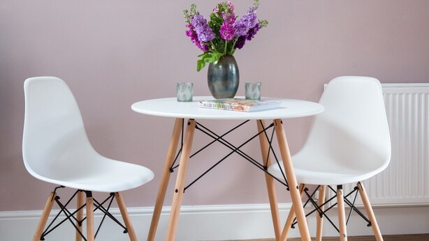 Une pièce avec deux chaises blanches, une petite table ronde et un vase rempli de fleurs violettes.