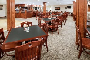 Comedor de hotel con varias mesas y sillas de madera, una zona de bar con estantes llenos de botellas y suelo alfombrado.