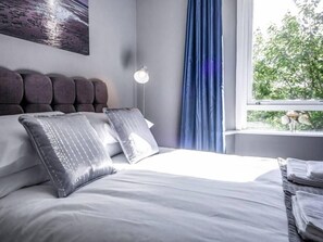 A neatly made bed with white linens and two decorative pillows, a modern headboard, a floor lamp, and a view of greenery through a window with blue curtains.