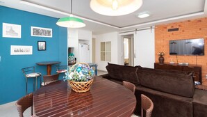 Un salon moderne avec un coin repas, comprenant un mur bleu orné de tableaux encadrés, une table à manger en bois avec un panier de fleurs, un canapé marron et un téléviseur fixé à un mur en briques.