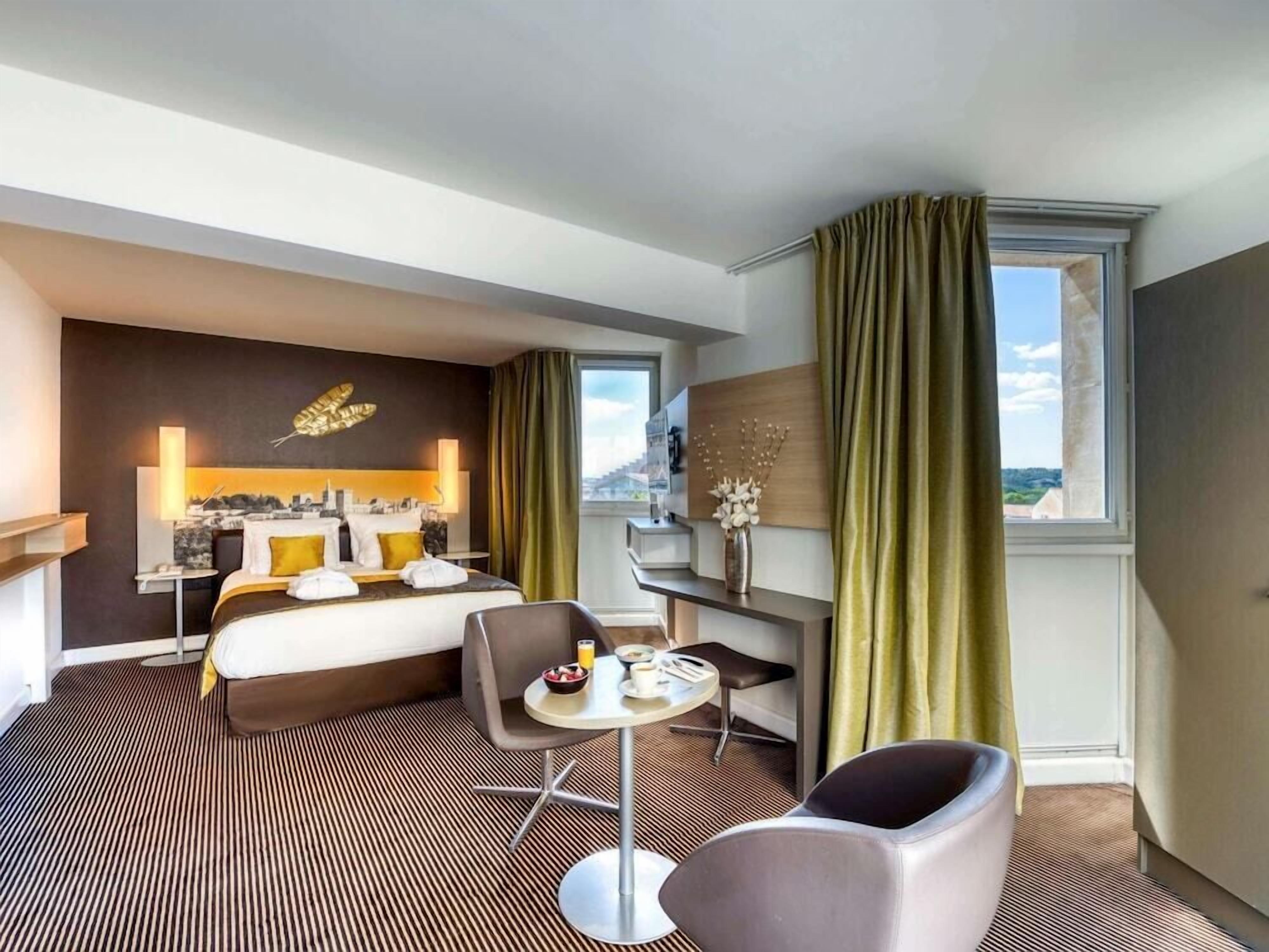 A hotel room with a bed, a desk, a chair, a small table with breakfast items, and a view of the outdoors.