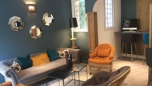 Una sala de estar moderna con un sofá gris, una mesa de centro negra y un sillón naranja. Hay un escritorio de madera con una computadora y una silla. La habitación tiene un ventanal grande y espejos decorativos en la pared.