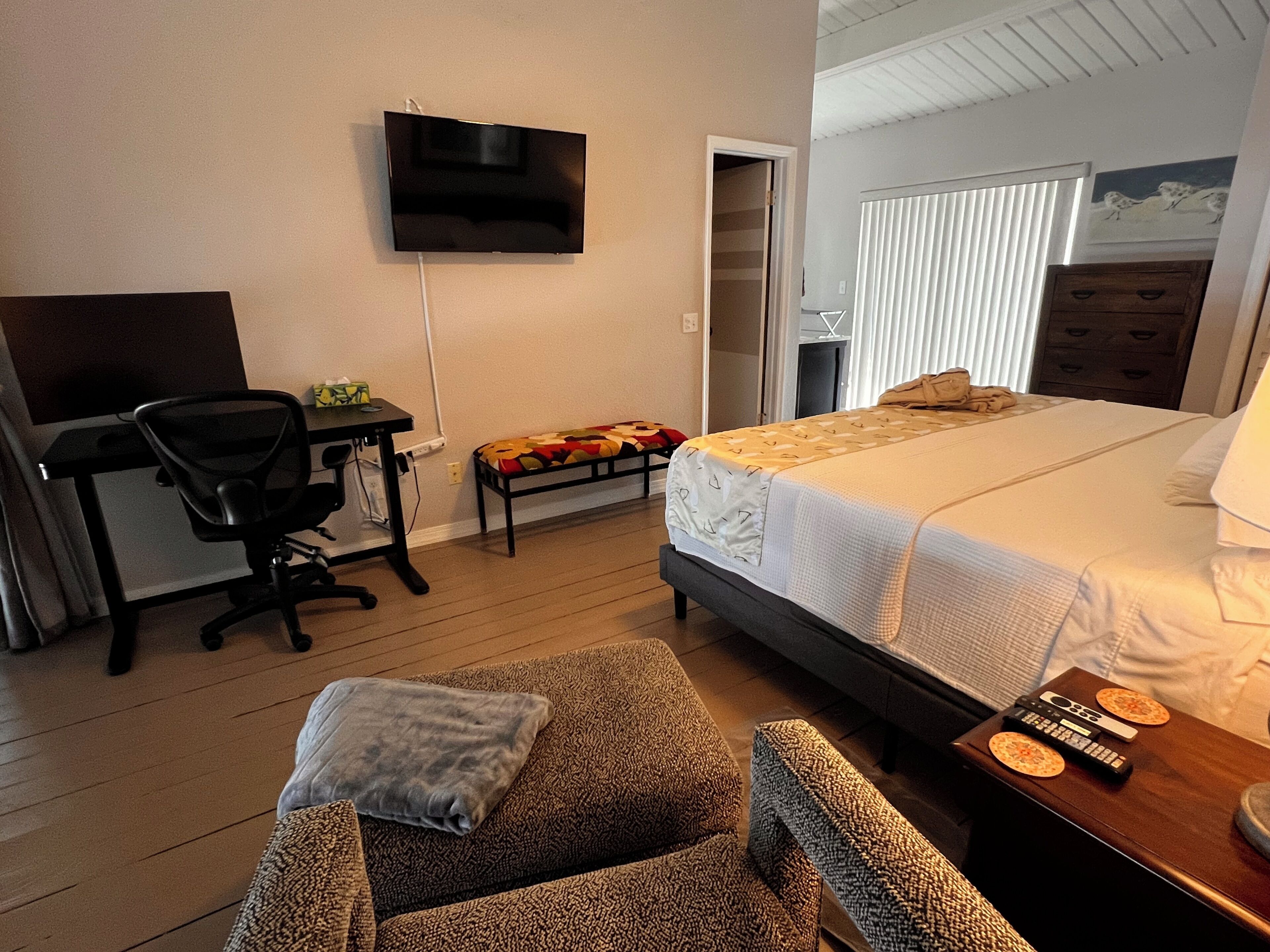 A bedroom with a bed, a desk, a chair, a TV, and a small table with a remote control.