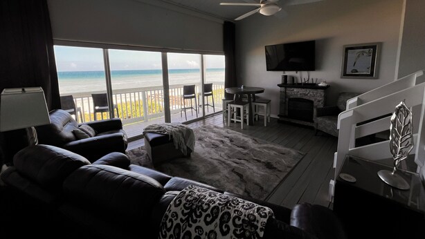 A living room with a view of the ocean, a ceiling fan, a fireplace, and a TV mounted on the wall.