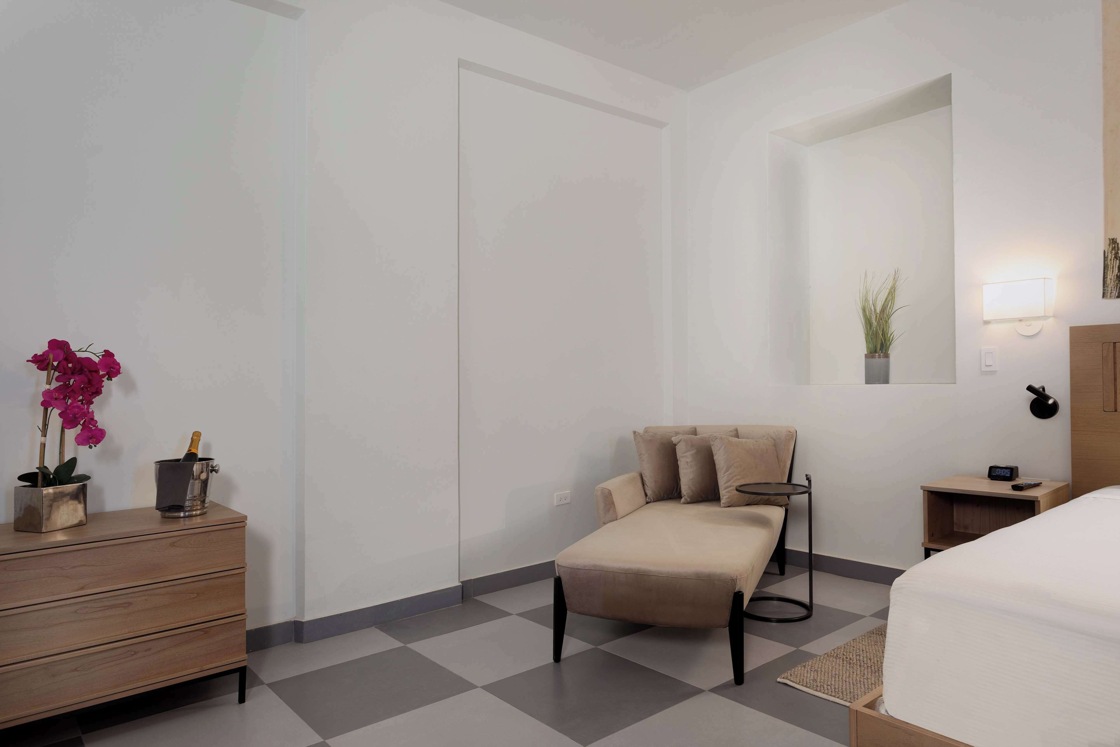 A modern hotel room with a checkered floor, a sofa, a wooden dresser, a bedside table with a lamp, and a mirror on the wall.