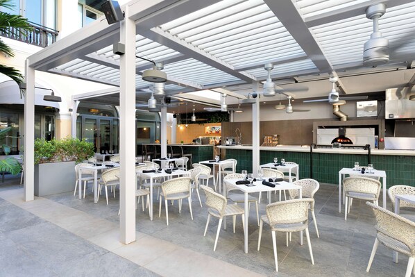 Un espacio de patio al aire libre con sillas y mesas de mimbre blanco, una zona de estar cubierta y una barra con horno para pizza.