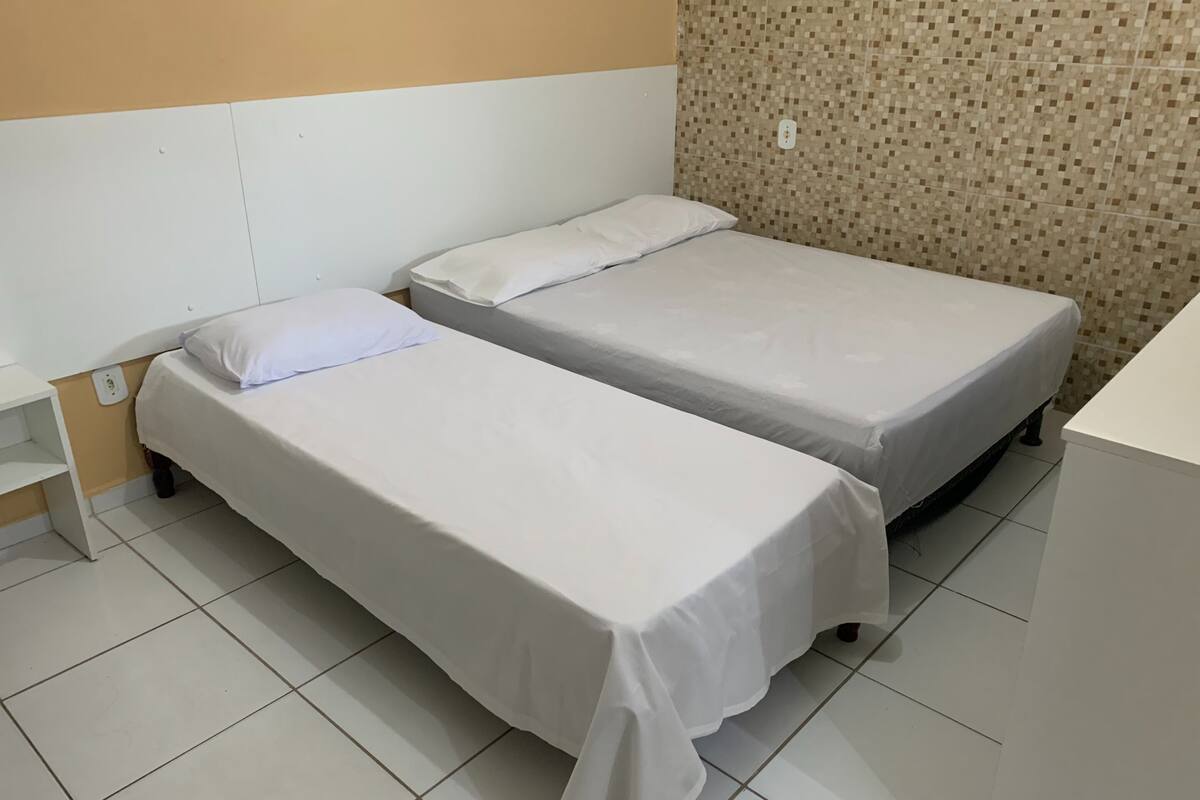 Quarto de hotel com duas camas de solteiro, uma pequena mesa de cabeceira e piso ladrilhado.