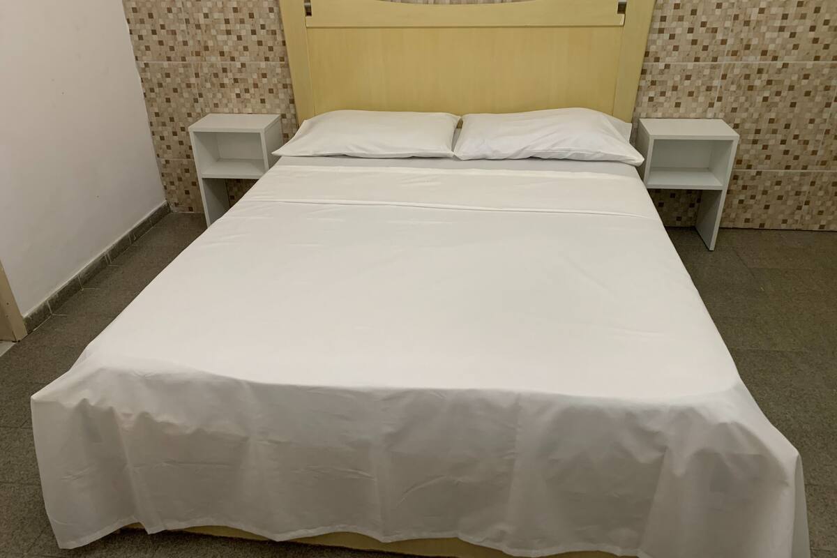 Cama bem arrumada com colcha branca, duas mesas de cabeceira e encosto.
