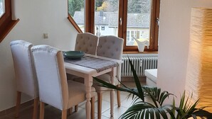 A dining area with a table and chairs, a clock on the wall, and a view of the outside through large windows. - Ferienwohnung Bärenhofweg am Titisee (Titisee)