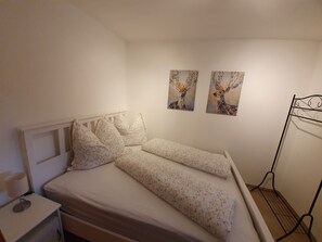 Ein Einzelbett mit floralem Bettbezug in einem Zimmer mit weißen Wänden. Zwei an der Wand befestigte Bilder von Hirschen. Ein Metallregal steht rechts an der Wand.