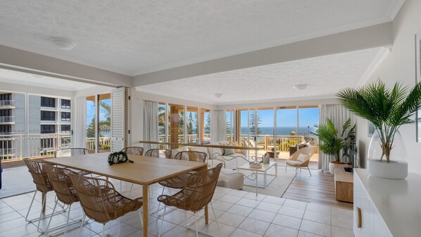 A spacious living area with a large table and chairs, a balcony with a view of the sea, and a large window providing natural light.