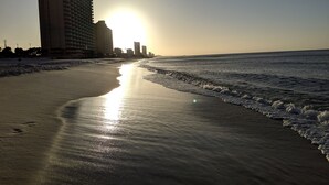 Une plage où le soleil se reflète sur le sable, une jetée au loin et un grand bâtiment d’hôtel.
