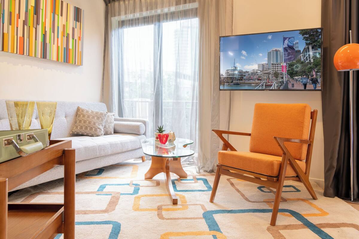 A modern living room with a sofa, a coffee table, and a chair. There is a large window with sheer curtains, a painting of a cityscape, and a colorful abstract wall art piece.
