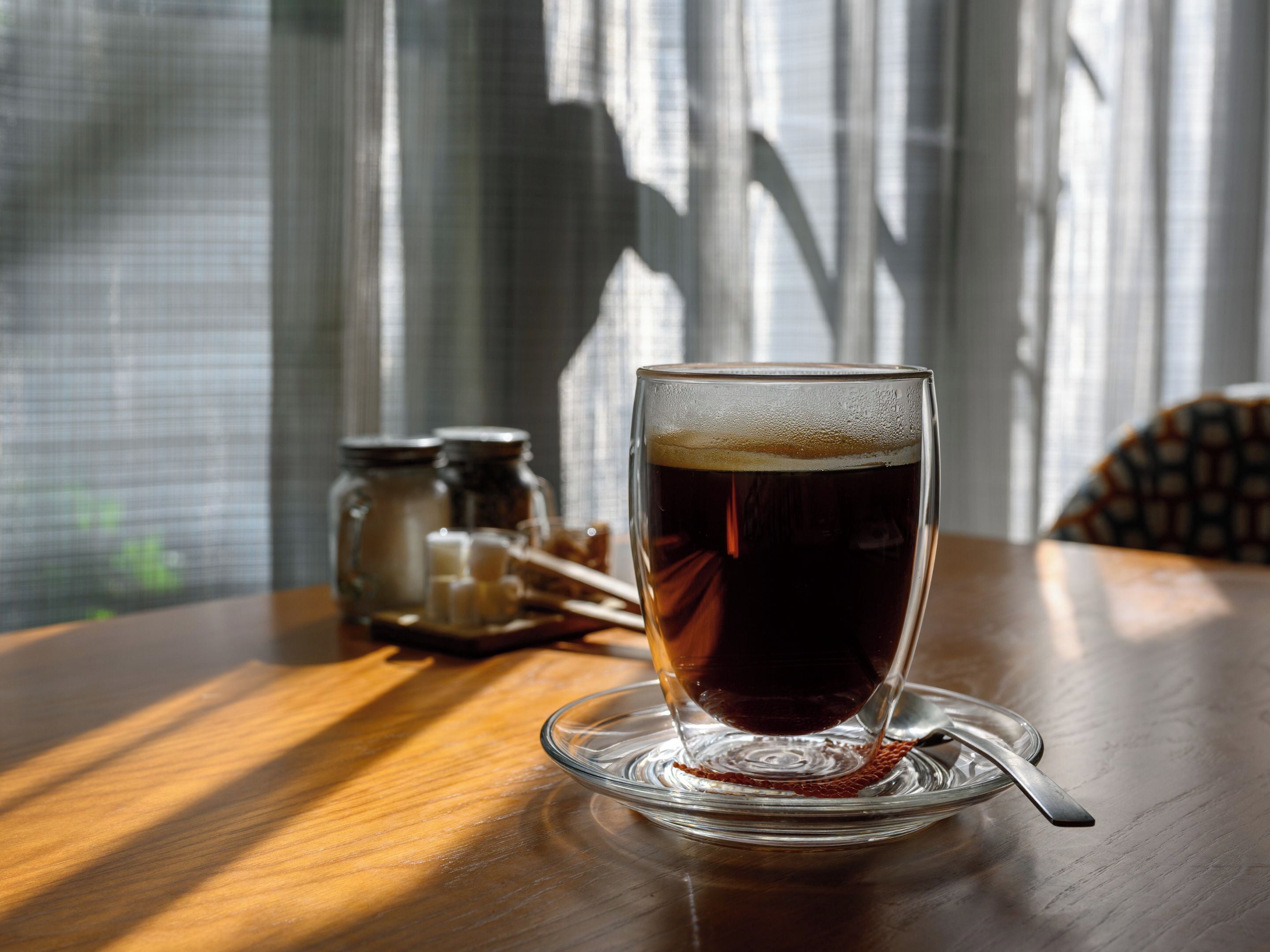 Un verre contenant un liquide sombre, posé sur une soucoupe en verre transparent, sur une table en bois.