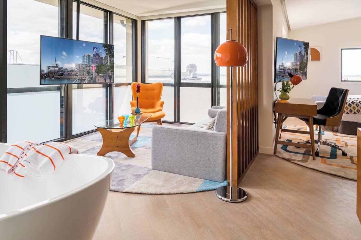A modern hotel room with a large window, a bathtub, a grey sofa, a wooden side table, and a desk with a chair.
