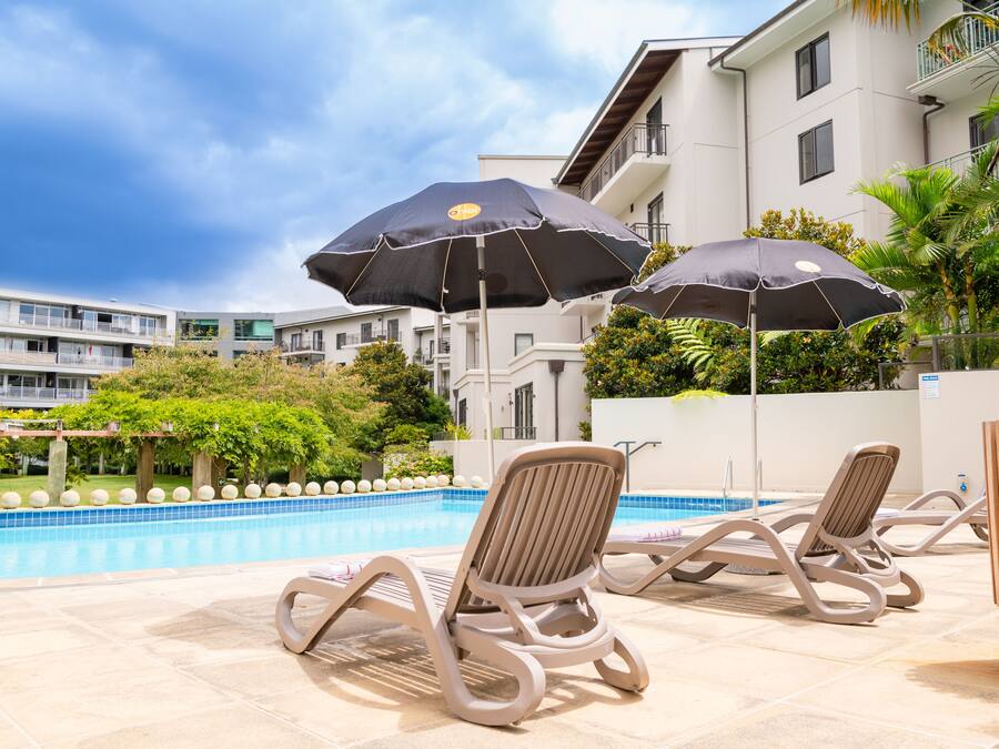 A poolside area with lounge chairs and umbrellas, a swimming pool, and a building with balconies.