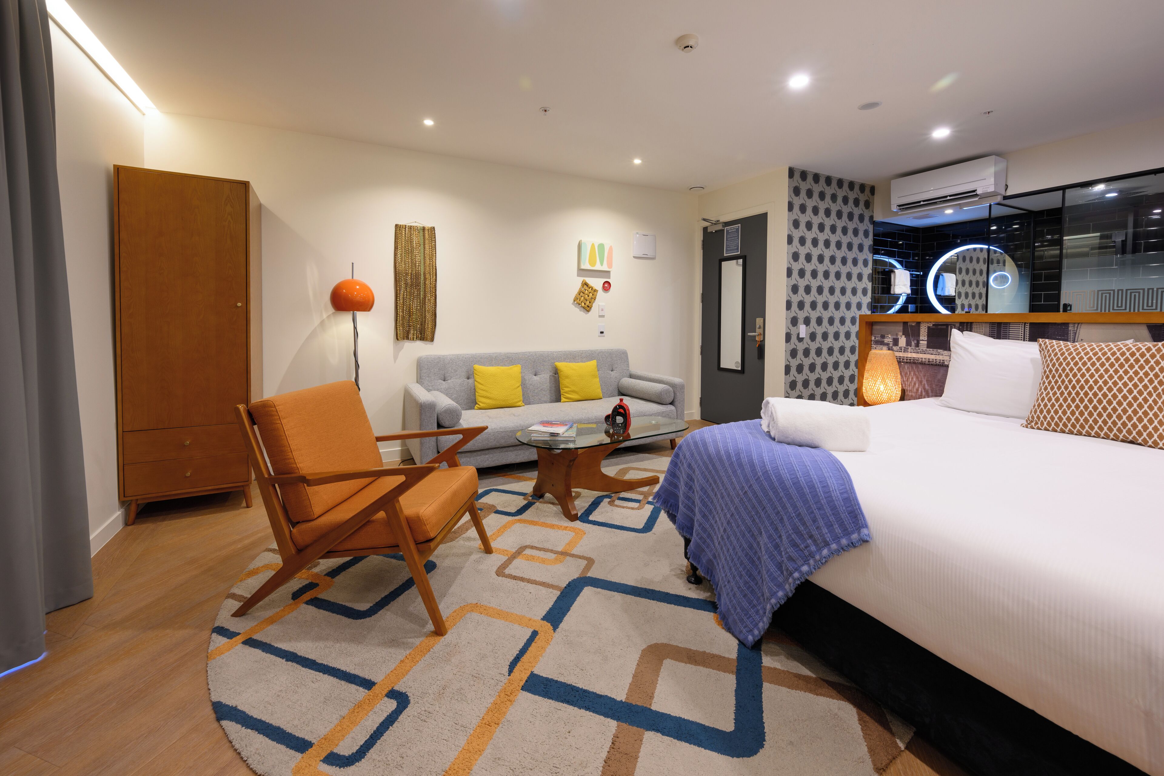 Habitación de hotel moderna con una cama grande, un sofá, una mesa de centro y una silla. Se ve una puerta, un aire acondicionado de pared y una ventana con vistas a la ciudad.