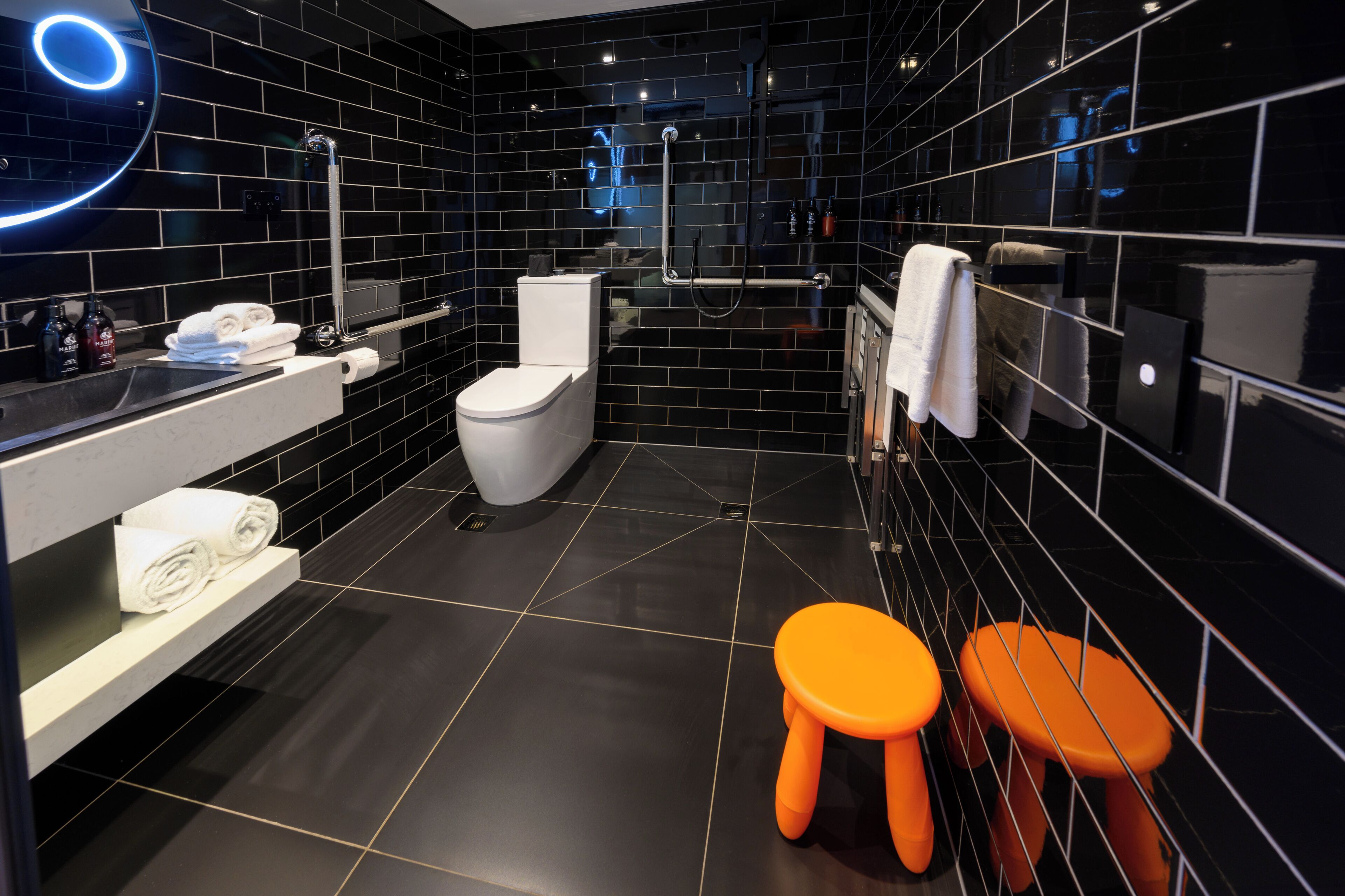 Un baño moderno con paredes de azulejos negros, un inodoro blanco, un lavabo con artículos de aseo y dos taburetes naranjas.