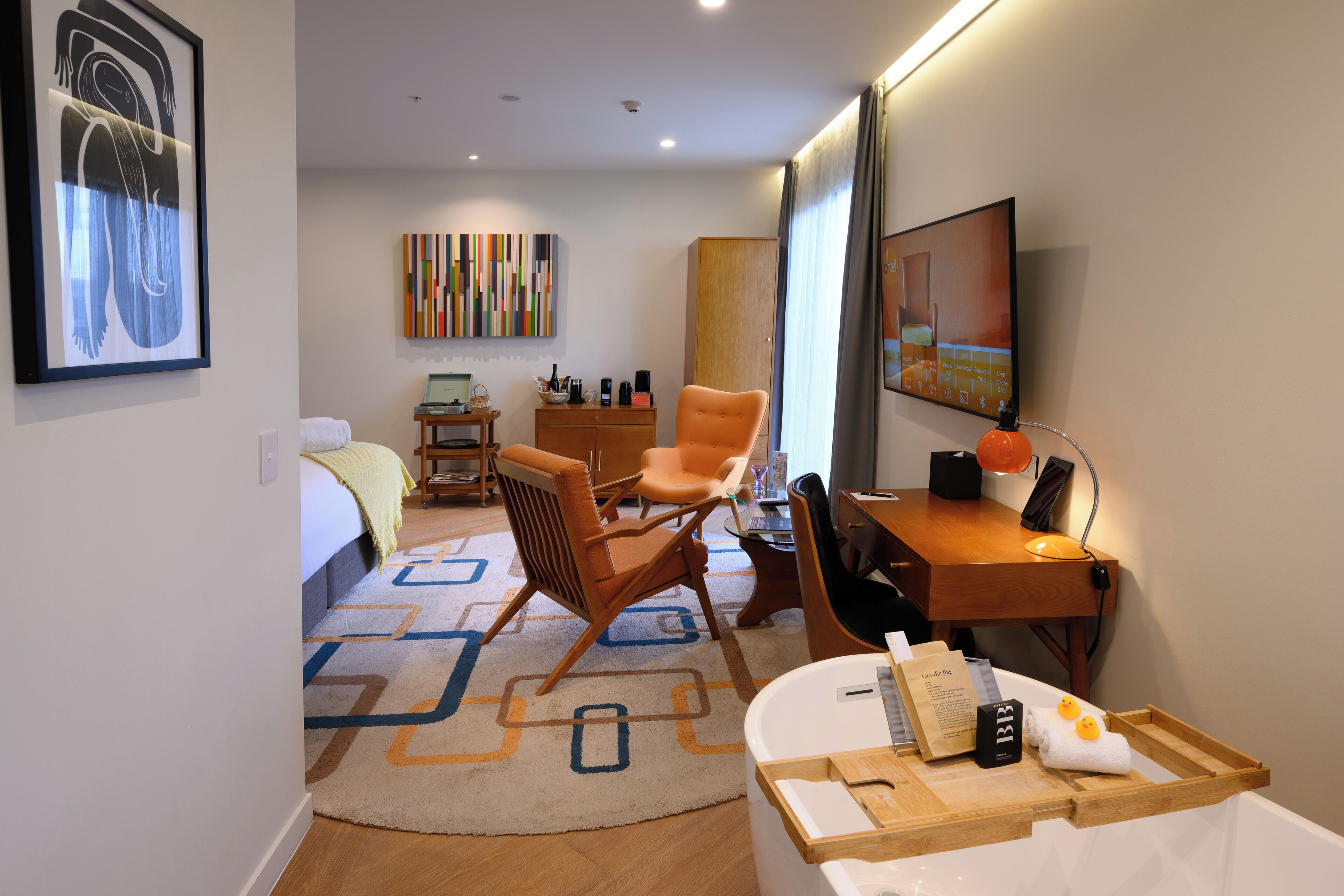 Hotelkamer met een bed, een bureau, een stoel, een bureaulamp, een televisie, een houten kast en een kleurrijk abstract schilderij aan de muur.