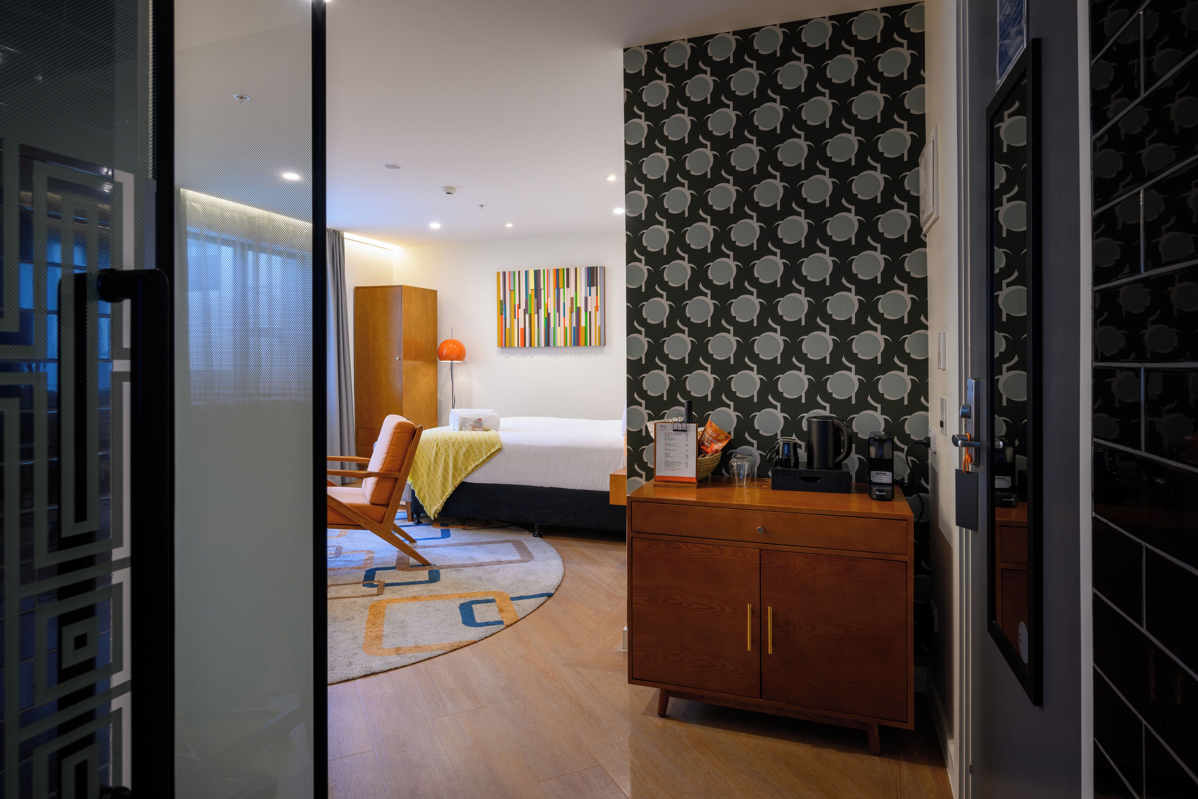 A hotel room with a bed, a desk with a chair, a wooden cabinet, and a patterned wall.
