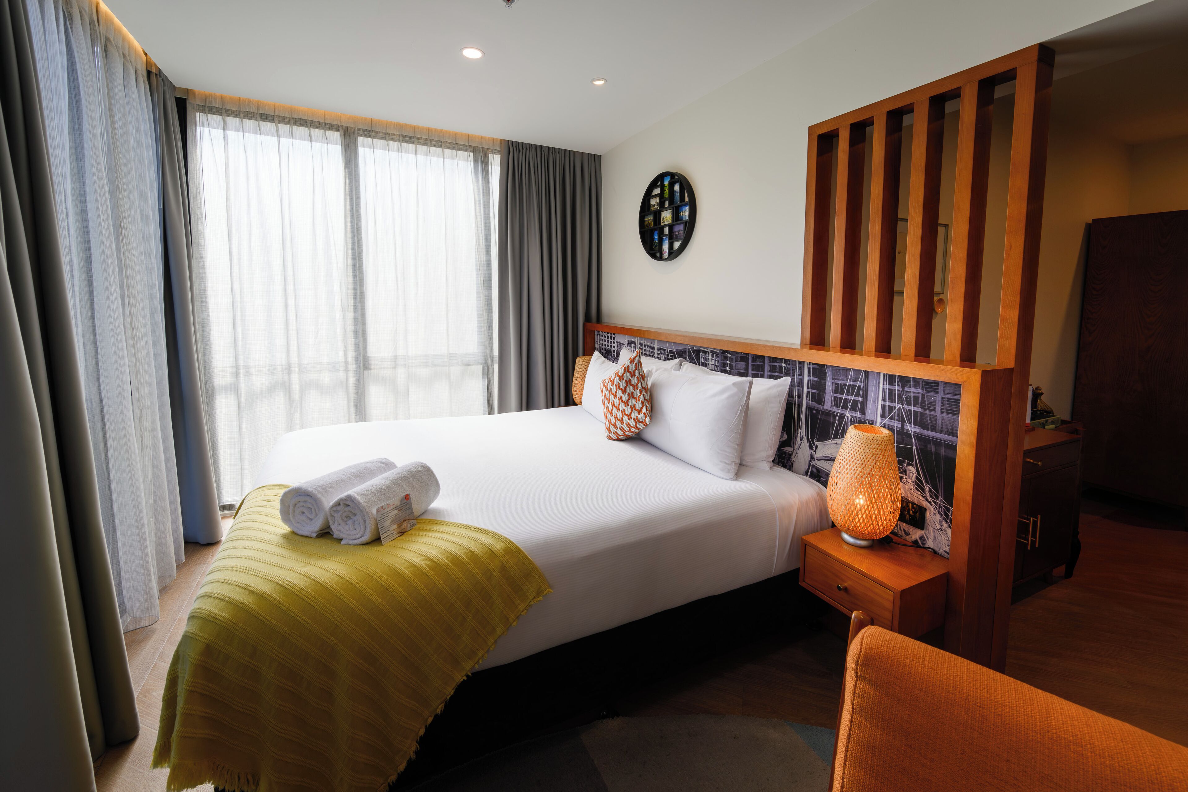 Une chambre d’hôtel avec un grand lit, une table de chevet avec une lampe, une tête de lit en bois et une vue sur la ville par la fenêtre.