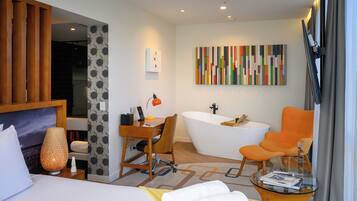 A modern hotel room with a bed, desk, chair, bathtub, and a glass coffee table.
