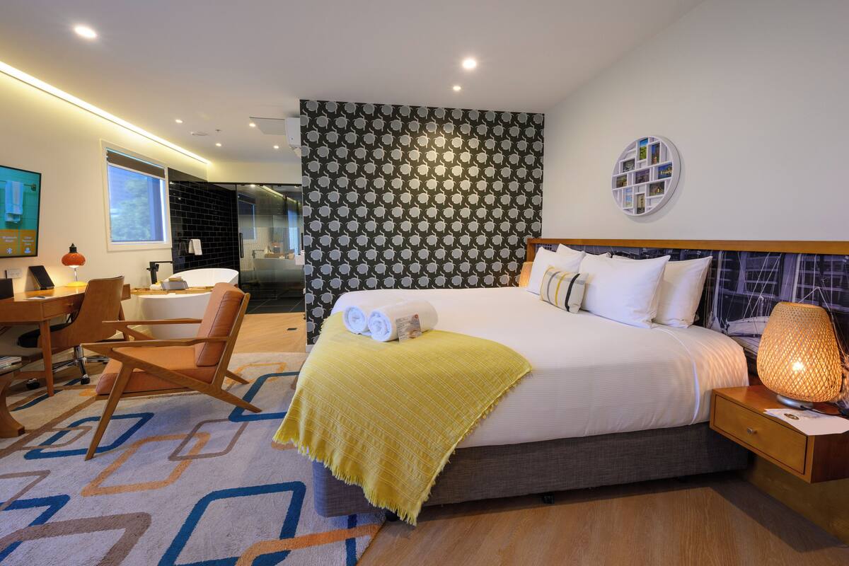 A modern hotel room with a large bed, a desk with a chair, a small table, and a wall-mounted TV.
