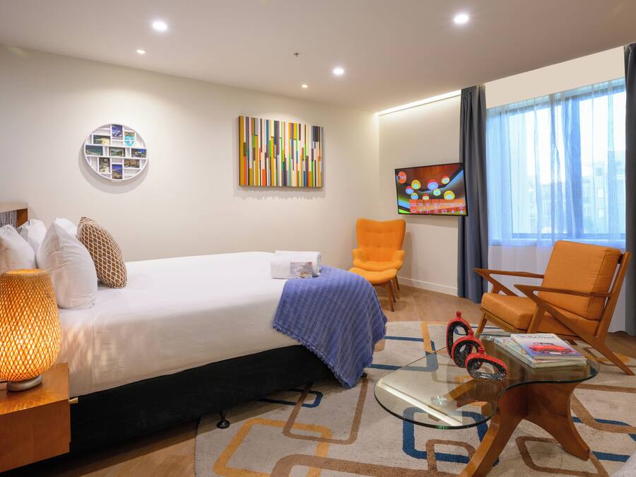 A modern hotel room with a neatly made bed, a glass coffee table, an orange armchair, and a large window with curtains.