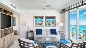 A modern living room with a sofa, two armchairs, a coffee table, and a TV. There are two paintings on the wall, a ceiling fan, and a large window with a view of the ocean.