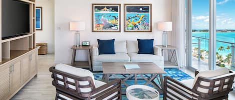 A modern living room with a sofa, two armchairs, a coffee table, and a TV. There are two paintings on the wall, a ceiling fan, and a large window with a view of the ocean.