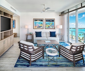 A modern living room with a sofa, two armchairs, a coffee table, and a TV. There are two paintings on the wall, a ceiling fan, and a large window with a view of the ocean.