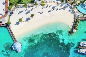 Vista aérea de un complejo turístico frente a una playa de arena blanca, con aguas turquesas cristalinas, un muelle y una zona de piscina con tumbonas.
