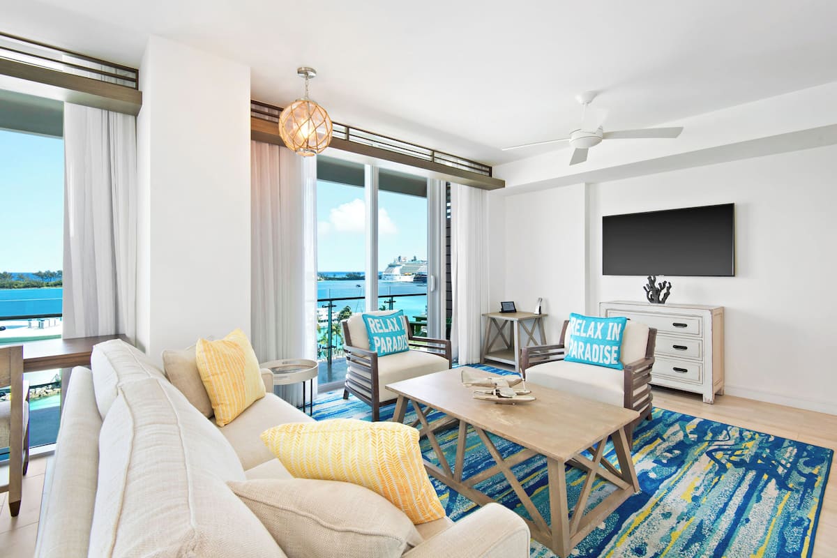 Ein modernes Wohnzimmer mit Sofa, Couchtisch und Fernseher. Ein Balkon bietet Blick auf das Meer und ein Kreuzfahrtschiff.