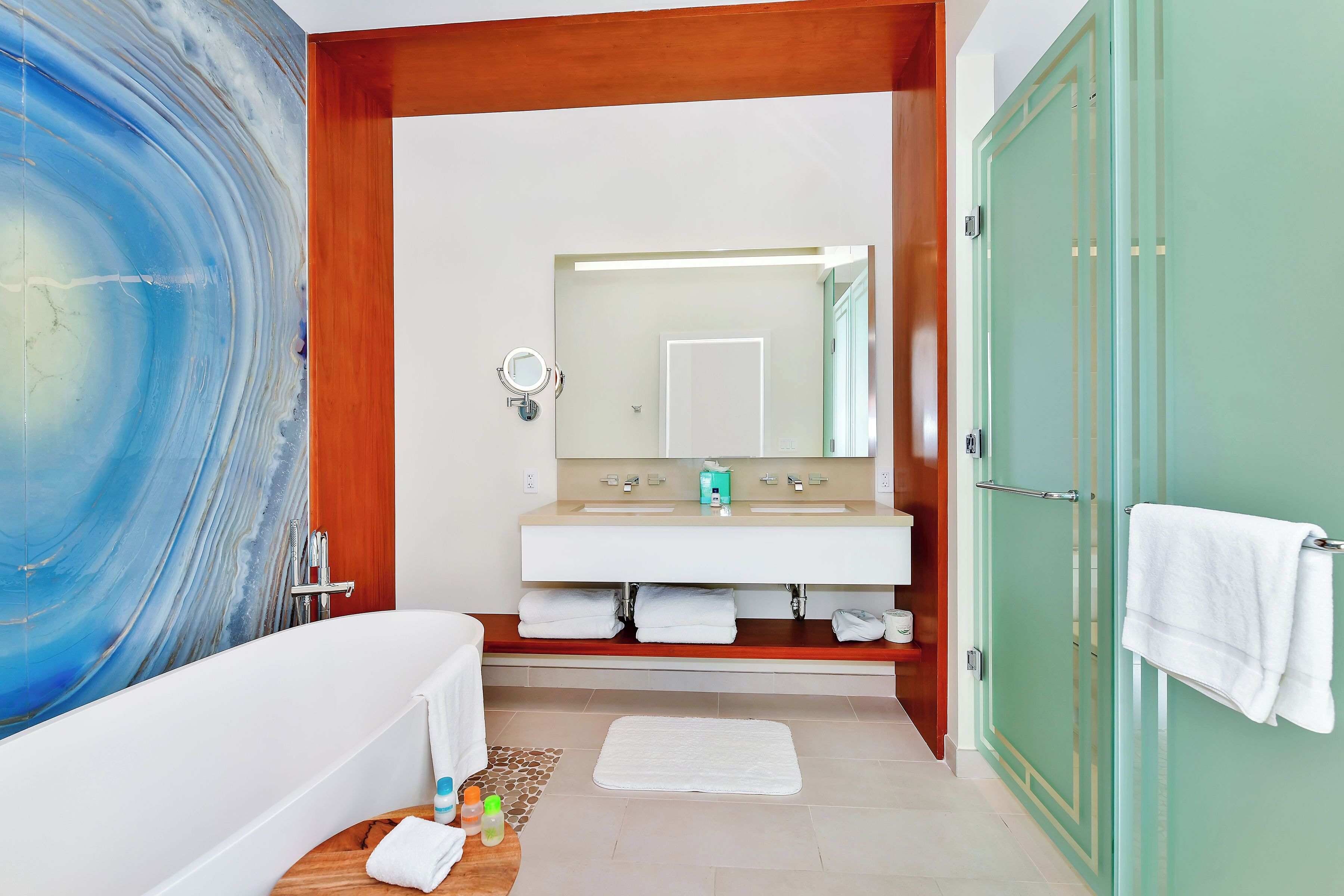 Une salle de bain avec un grand tableau abstrait bleu, une baignoire blanche, un meuble-lavabo en bois avec un miroir et une porte verte.