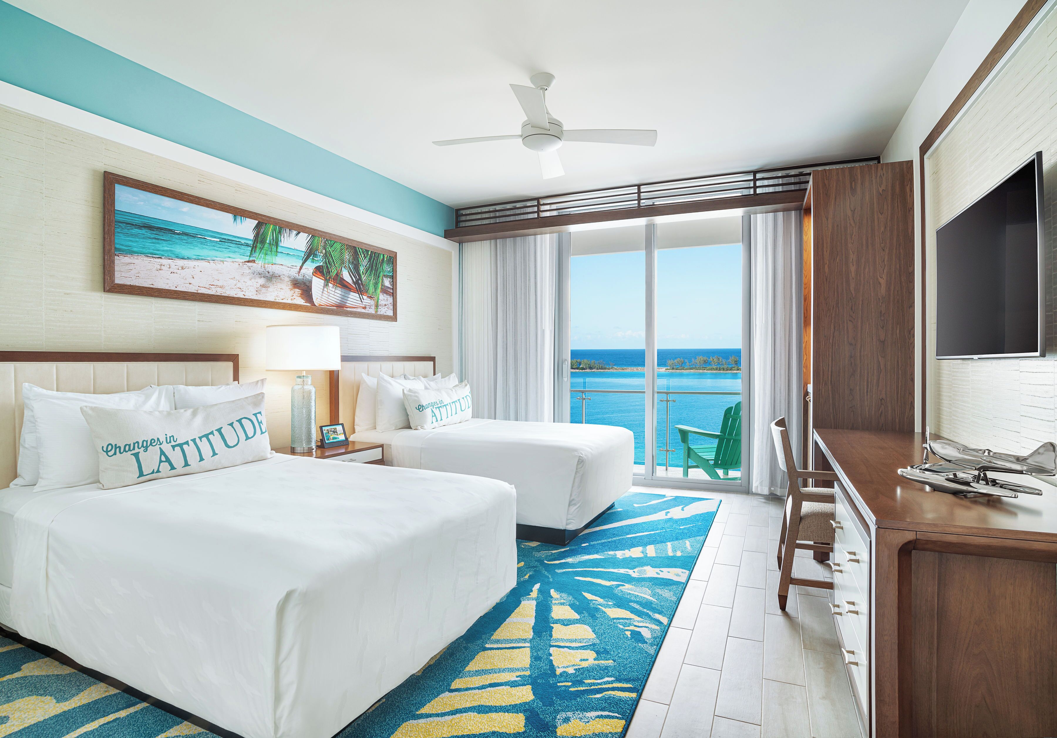 A hotel room with two beds, a ceiling fan, a large window with a view of the ocean, a flat-screen TV, and a wooden desk.