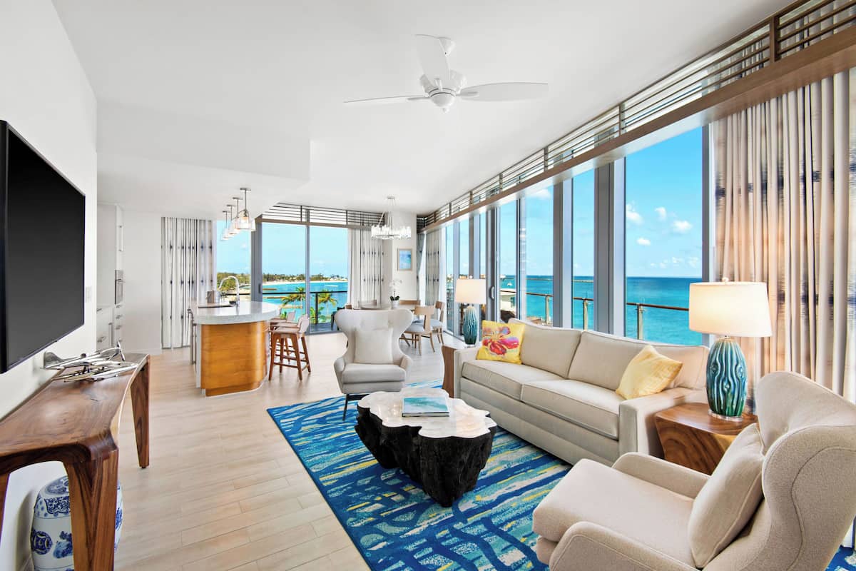 Ein geräumiges Wohnzimmer mit großem Fenster zum Meer, Deckenventilator und einer modernen Küchenzeile.